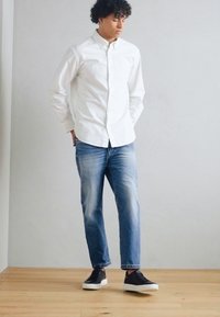 Chemise blanche à boutons, jean en denim bleu, baskets noires avec semelles blanches ; debout sur un sol en bois contre un mur gris clair.