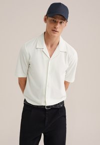 Camisa blanca de manga corta con un patrón texturizado y un cuello tipo revere, combinada con pantalones negros y un cinturón trenzado negro.