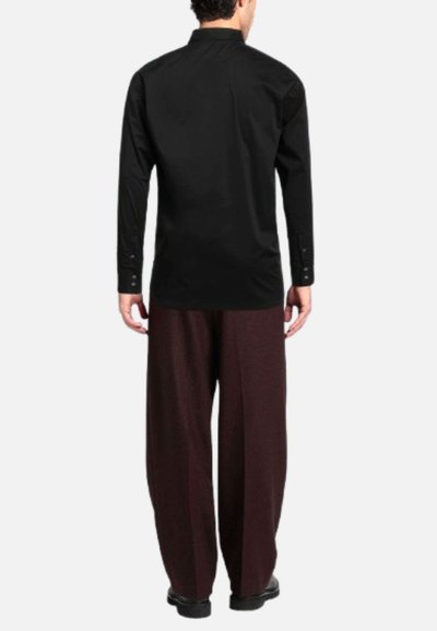 Uomo in piedi di spalle, indossando una camicia nera a maniche lunghe e pantaloni larghi marrone scuro con scarpe nere su uno sfondo semplice.