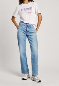 Jeans de mezclilla azul claro de corte recto, combinados con una camiseta gráfica blanca que lleva "LONDRES". Zapatillas deportivas grises completan el conjunto.