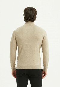 Pull col roulé beige avec un tricot texturé, une silhouette ajustée, et des poignets et un ourlet côtelés, associé à un pantalon noir.