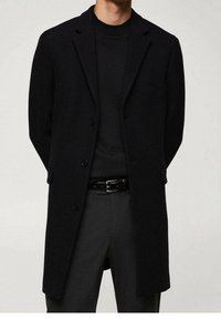 Manteau en laine noire avec un col à revers cranté, trois boutons et des poches latérales. Porté sur un pull noir, associé à un pantalon sombre à motifs.