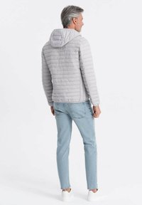 Lys grå pufferjakke med vandret syninger, fasthængt hætte og elastiske manchetter, parret med lyseblå jeans og hvide sneakers.