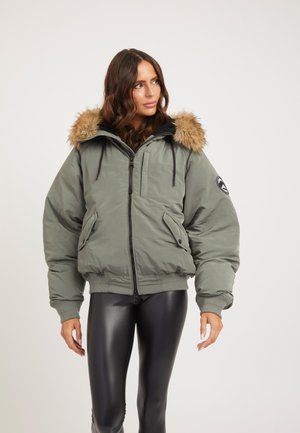 Mujer con chaqueta acolchada verde con capucha con ribete de piel y leggings negros de piel sintética, de pie frente a un fondo liso.
