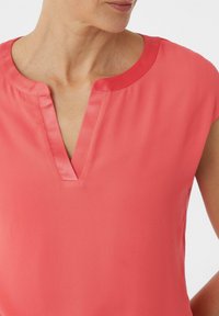 Koralowa bluzka z okrągłym dekoltem, z dziurką w kształcie klucza i subtelnym satynowym wykończeniem. Krótkie rękawy typu cap i gładka faktura.