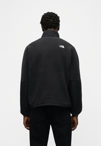 Άνδρας με μαύρο φλις μπουφάν με το λογότυπο The North Face στο πάνω μέρος της πλάτης, στέκεται μπροστά σε απλό φωτεινό φόντο, γυρισμένος προς τα πίσω.