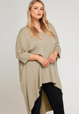 Beige, übergroßes Oberteil mit Rundhalsausschnitt und kurzen Ärmeln. Der Saum ist asymmetrisch und schafft eine fließende Silhouette. Kombiniert mit schwarzen Hosen.