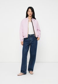 Jeune femme portant une veste bomber rose clair, un haut blanc, un jean bleu à coupe large et des ballerines rose pâle, se tenant devant un fond blanc.