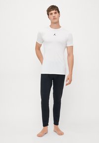 Un homme se tient pieds nus, portant une chemise blanche ajustée à manches courtes avec un logo noir et un pantalon noir slim, devant un fond blanc uni.