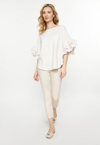 Blusa color beige chiaro con maniche arricciate e un delicato motivo a foglie, abbinata a pantaloni aderenti color crema e ballerine nude, su uno sfondo neutro.