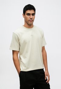 Šviesiai smėlio spalvos medvilninė marškinėliai trumpomis rankovėmis, apvalia iškirpte ir subtiliu „BOSS“ logotipu ant krūtinės; dėvimi su tamsiomis kelnėmis.