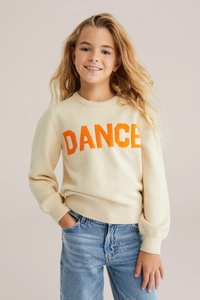 Krämfärgad stickad tröja med djärv orange "DANCE" text, med ribbade ärmslut och nederkant. Tillsammans med ljusblå jeans prydda med små dekorationer.