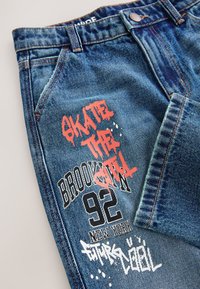 Blue denim jeans with graffiti-style text in red, black, and white reading "SKATE THE CITY," "BROOKLYN 92," and "NEW YORK."