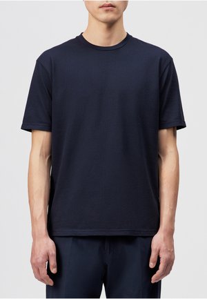 Hombre con camiseta lisa de manga corta y cuello redondo de color azul marino oscuro y pantalones oscuros, de pie contra un fondo claro y liso.