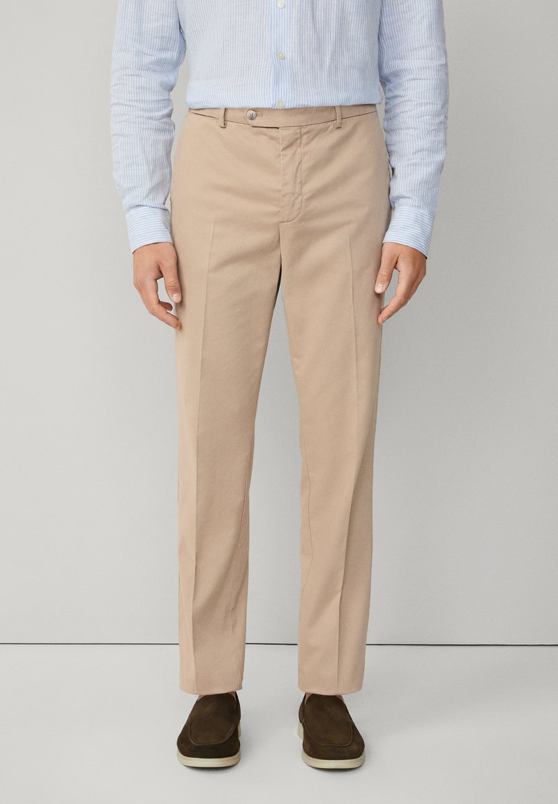 Uomo che indossa pantaloni beige su misura, camicia azzurro chiaro a righe verticali e scarpe scamosciate marrone scuro senza lacci, in piedi davanti a uno sfondo neutro.