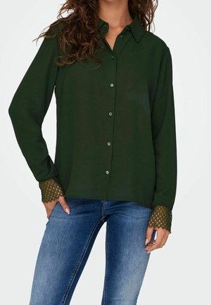 Femme portant un chemisier vert foncé boutonné avec des poignets en dentelle, associé à un jean bleu en denim, debout devant un fond uni.