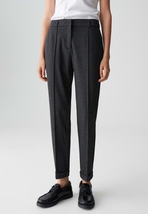 Zuschnitt dunkelgraue maßgeschneiderte Hose mit einer Frontfalte, mit umgeschlagenen Säumen, getragen mit schwarzen Schnürschuhen und einem weißen, hineingesteckten Oberteil.