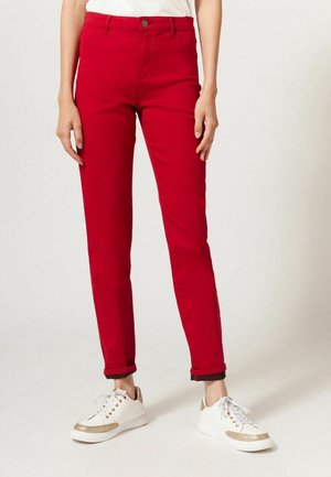Pantalon slim rouge vif avec ourlets retroussés, porté avec des baskets blanches aux accents dorés et un haut blanc rentré.