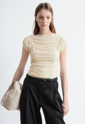 Jeune femme aux cheveux bruns raides portant un haut crème froncé, un pantalon noir, tenant un sac à main beige sur fond blanc.