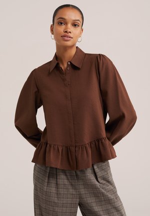 Femme portant un chemisier marron à manches longues avec un ourlet volanté et un pantalon taille haute à carreaux, debout devant un fond beige uni.