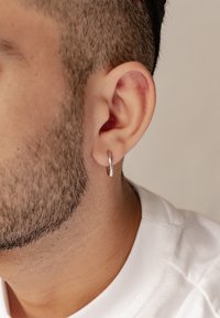 Silberne Creolen, die an einem durchstochenen Ohr befestigt sind. Das Ohr zeigt kurzen Gesichtshaarschmuck, mit einem glatten Hals und einem sichtbaren weißen Hemd.