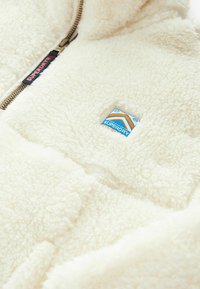 Veste en polaire de couleur crème avec fermeture éclair, tissu texturé et un écusson de logo Superdry bleu et orange sur le devant.