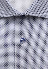 Col de chemise bleu et blanc avec un motif géométrique en losange et un bouton bleu au centre sous le col.