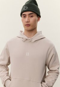 Junger Mann, der einen beigefarbenen Hoodie mit "II" auf der Brust und eine dunkelgrüne Mütze mit einem weißen gestickten Motiv trägt.