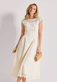 Weißes Midi- Kleid aus Spitze mit floralen Ausschnitten, kurzen Ärmeln und gewelltem Saum. Kombiniert mit einer geflochtenen Handtasche für zusätzlichen Stil.