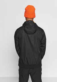 Sort hættejakke med en glat overflade og elastiske manchetter, båret med en orange strikhue, set fra ryggen.