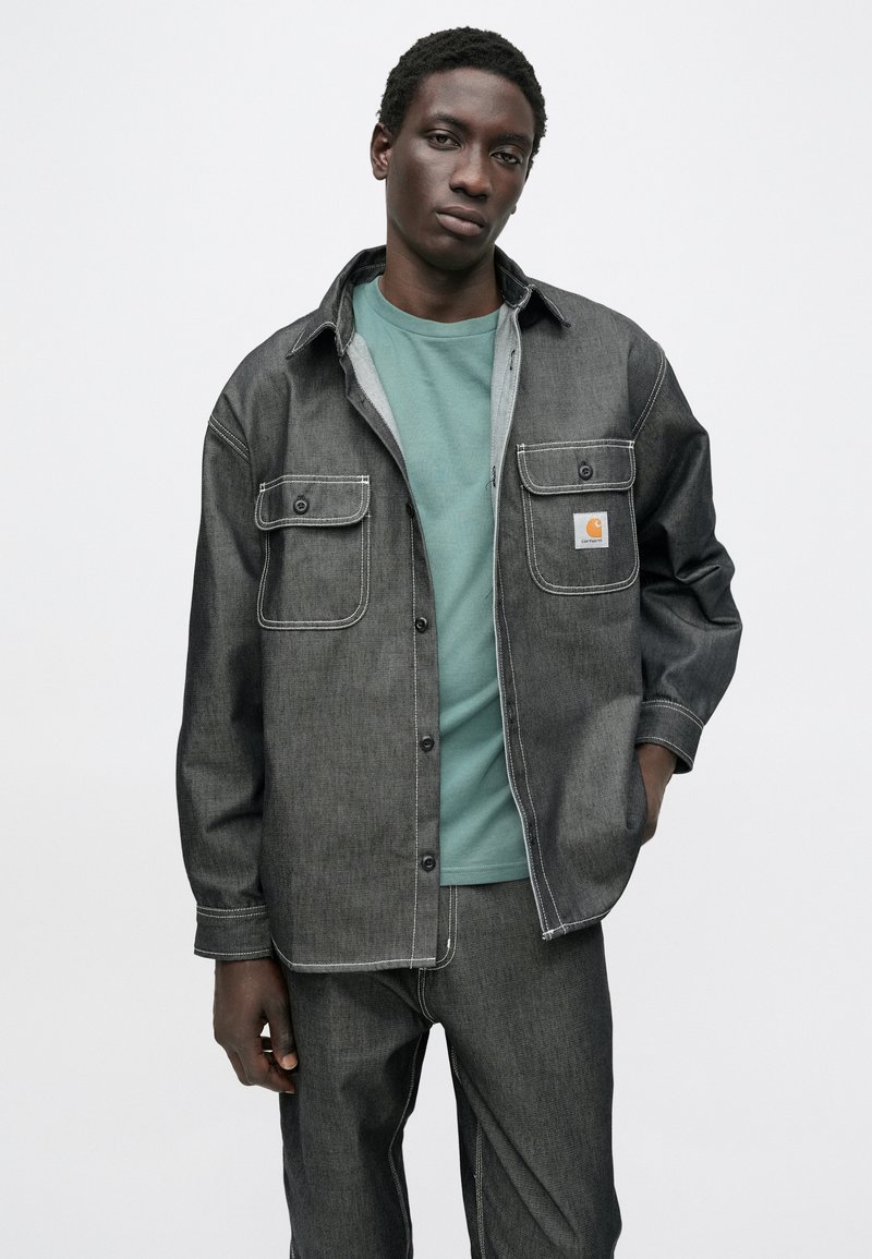 Giacca di denim scuro con due tasche frontali, cuciture azzurro chiaro e logo Carhartt. Indossata sopra una maglietta turchese. Sfondo neutro.