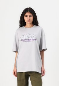 T-shirt gris surdimensionné avec un graphique brillant « ALPHA STUDIO EDITION » en violet et blanc. Manches courtes et col rond.