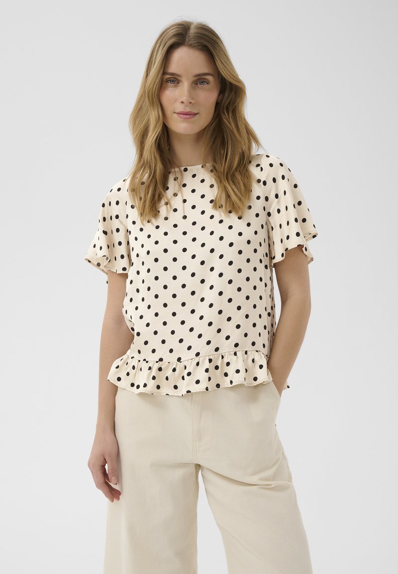 Femme aux longs cheveux ondulés portant une blouse crème à pois noirs avec un ourlet à volants, associée à un pantalon beige clair, debout devant un fond uni.