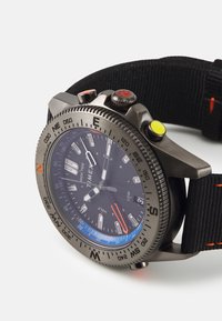Montre Timex avec un boîtier rond en acier inoxydable gris foncé, un bracelet en nylon noir, un cadran noir, des index lumineux et des boutons colorés pour les fonctions.