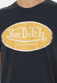 T-shirt en coton bleu marine avec un graphique ovale jaune usé portant le texte "Von Dutch" et "CALIFORNIA" en blanc. Manches courtes, col rond.