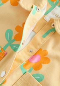 Camasa cu guler din material galben, cu modele mari, colorate de flori și frunze în verde, portocaliu și roz. Se observă închiderile cu capse.