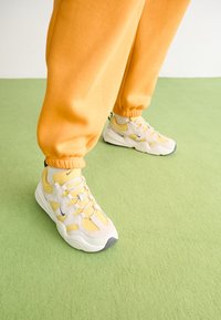 Ljust grå och gula sneakers med mesh- och mockamaterial, med tjocka sulor och framträdande snören, kombinerade med orangea joggers.