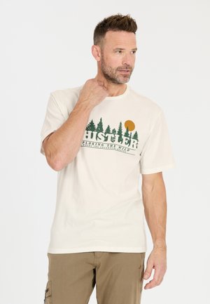 Mies, jolla on lyhyet hiukset ja parta, koskettaa kaulaansa. Hänellä on päällä beige t-paita, jossa on vihreitä puita ja oranssi aurinko, sekä ruskeat housut. Taustana on valkoinen väri.