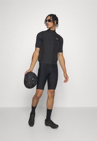 Schwarzes Radtrikot mit kurzen Ärmeln und Reißverschluss vorne, kombiniert mit schwarzen gepolsterten Shorts. Die Accessoires umfassen schwarze Radschuhe und einen Helm.
