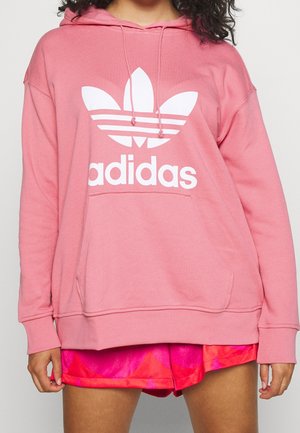 Person iført en løs pink Adidas-hoodie med en stor hvid logo og lyse, mønstrede pink og røde shorts, stående mod en hvid baggrund.