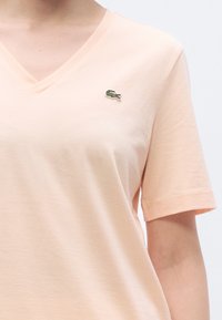 Ροδακινί βαμβακερό μπλουζάκι με V-λαιμόκοψη και κοντά μανίκια. Διαθέτει μικρό πράσινο κεντημένο λογότυπο κροκόδειλου στο στήθος.