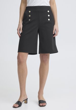 Žena v čiernych vysokých krátkych nohaviciach po kolená so šiestimi kovovými gombíkmi a čiernych sandáloch na podpätku s otvorenou špičkou.