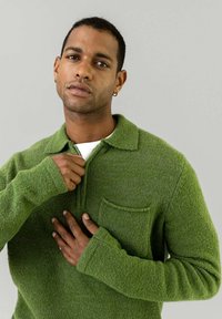 Grøn zip-up sweater lavet af tekstureret stof med krave, forbens lomme og lange ærmer, båret af en person med kort hår.