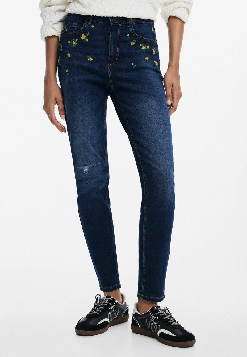 Tummansinisiä farkkuja, joissa on keltaista ja vihreää kukkakirjontaa edessä, slim fit -malli, jossa on hieman kulumista yhdessä polvessa. Mustat lenkkarit.