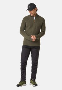 Olive green knitted sweater with a zip collar, paired with black pants featuring zippered pockets and green athletic shoes. Black cap worn.