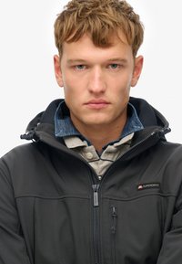 Zwarte jas met capuchon, gemaakt van gladde stof, met een ritssluiting aan de voorkant en een logo op de borst, gedragen over een lichtgrijs en denim overhemd.