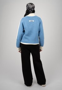 Personne portant un pull bleu avec des poignets blancs et un pantalon noir à jambes larges, vue de dos, avec un foulard noir à motifs noué à l'arrière.