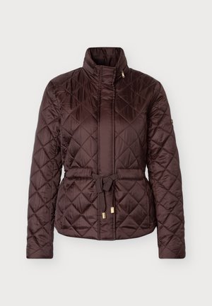 Femme portant une veste matelassée marron foncé avec ceinture, un jean bleu droit et des chaussures noires à bout pointu, debout devant un fond clair.