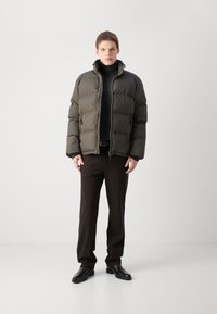 Jachetă puffer verde închis, cu guler înalt, închidere cu fermoar și textură matlasată, asortată cu pantaloni închiși la culoare și pantofi negri.