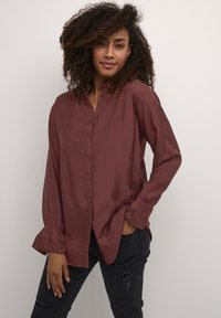 Camicia button-up bordeaux con una texture morbida e setosa, maniche lunghe con polsini arricciati e una vestibilità rilassata, indossata con jeans neri.